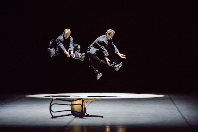 Two men jump with their legs outstretched into the air. Chairs lay on the ground. 